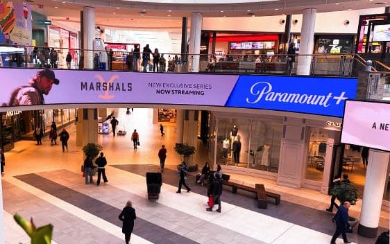 Multi-level shopping centre interior featuring a large-format advertising display above a central walkway, with shoppers throughout the space.