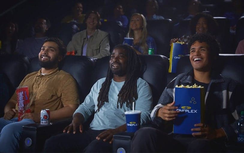 Group of moviegoers seated in Cineplex reclining chairs enjoying popcorn and drinks during a film
