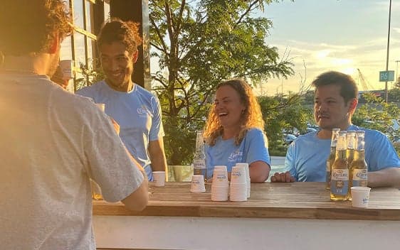 People smiling and serving drinks at an outdoor branded activation booth during sunset