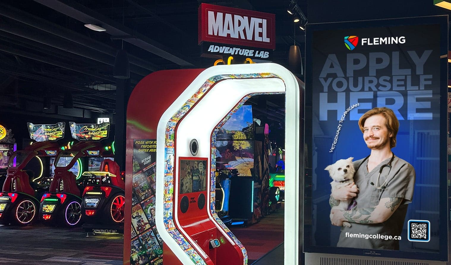 Digital screen at Playdium Whitby showing Fleming College advertisement beside arcade games