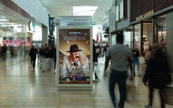 Mall shoppers walking past a large digital ad featuring a man in a hat and suit