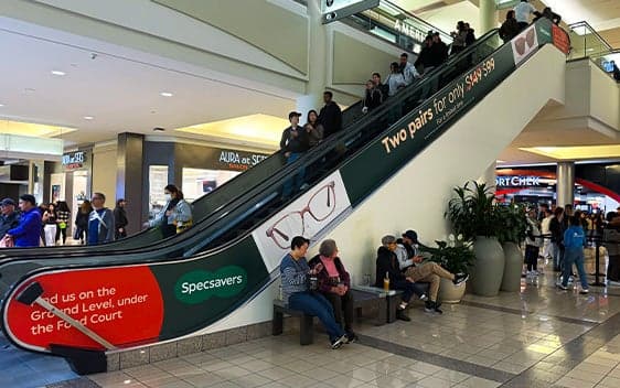 Escalator in busy mall wrapped with Specsavers advertising about eyewear deals